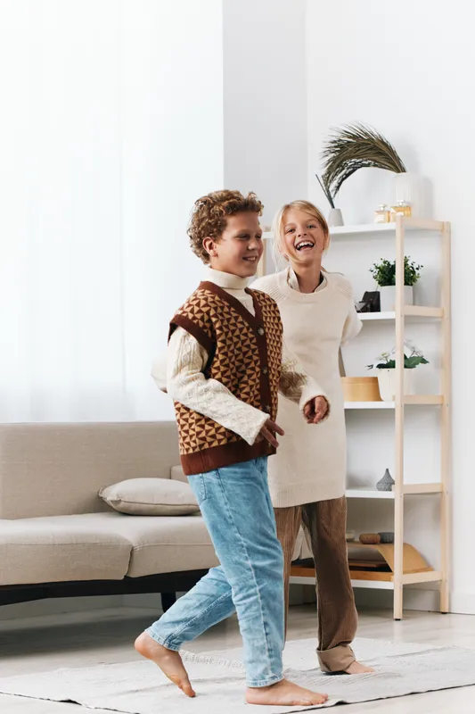 Two cheerful children laugh and play indoors, wearing cozy knit sweaters and soft trousers as they chase each other across a bright living room with a plush rug.
