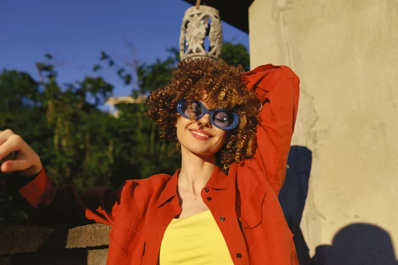 A stylish woman enjoys a sunny outdoor moment in a bright red jacket and yellow top, posing with a carefree smile. A lifestyle portrait that showcases confidence, fashion, and vibrant energy