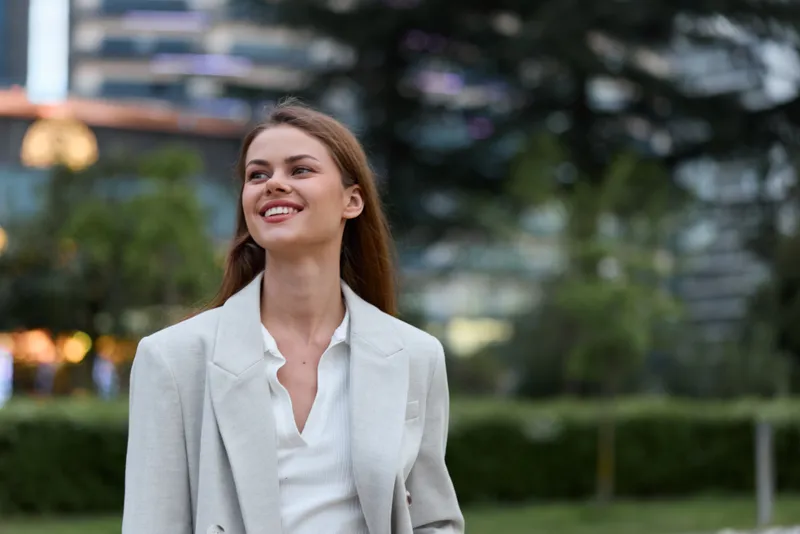 Confident businesswoman smiling outdoors in a stylish blazer, exuding professionalism with a vibrant urban background, showcasing modern corporate fashion.