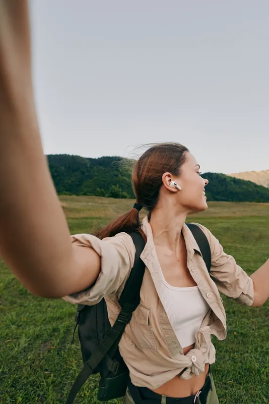 A woman with a backpack raises her arms in a bright outdoor field, smiling, embracing nature and freedom during a hiking adventure.