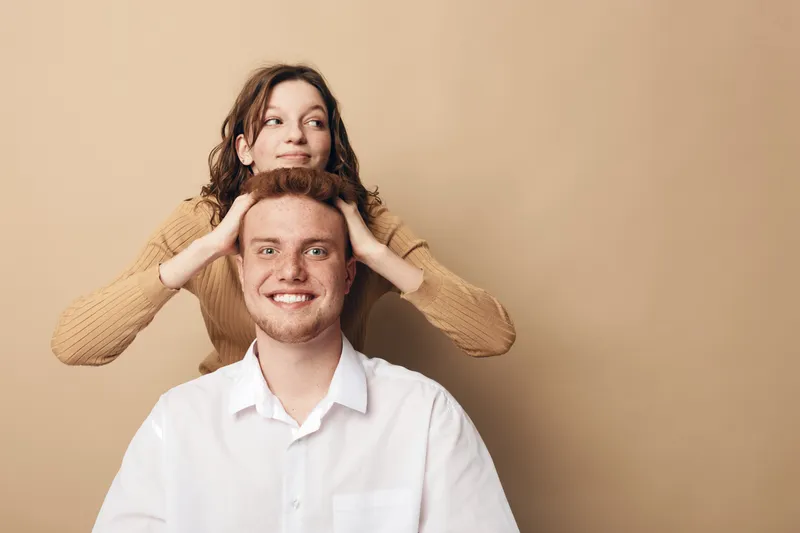 Smiling young man in white shirt sits while woman in beige sweater stands behind with hands on his head. Studio portrait on beige background. Concept of friendship, happiness, and casual lifestyle.