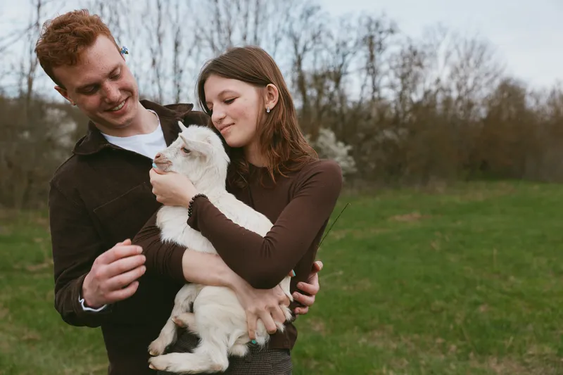 couple outdoors goat kid lifestyle nature field, warm candid moment, relaxed posture, sunlit colors, gentle breeze, lush greens, close bond, wholesome countryside vibe