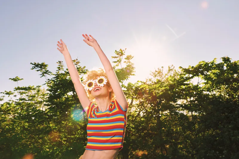 Joyful woman in a striped top with flower sunglasses raises arms outdoors, showcasing a carefree summer lifestyle with retro film color against a sunny park backdrop.