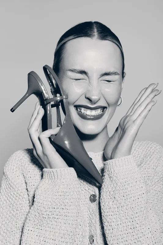 Smiling white woman with light skin holds high heel shoe near face, eyes closed, wearing knitted sweater. Concept of fashion and joyful emotion.