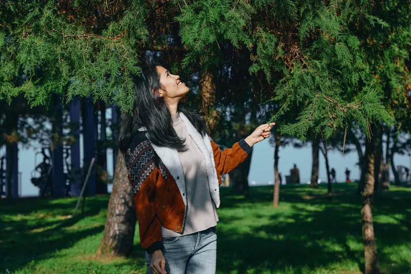 Woman enjoying nature under trees in a sunny park, looking up with outstretched hand, autumn jacket, scarf, and casual attire, capturing outdoor scenery, fresh air, and tranquil mood