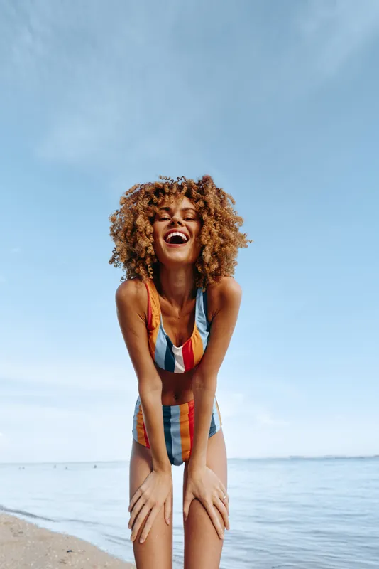 Woman curly hair beach ocean swimsuit summer laughter sunlight vacation outdoors laughing while sitting on sandy shore under blue sky, calm sea horizon, relaxed posture, summer trip joy