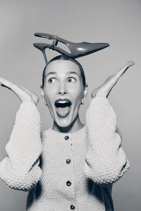 Young white woman with light skin wearing a knitted sweater balances a high heel shoe on her head and shows surprised expression in studio. Concept of fun and fashion.