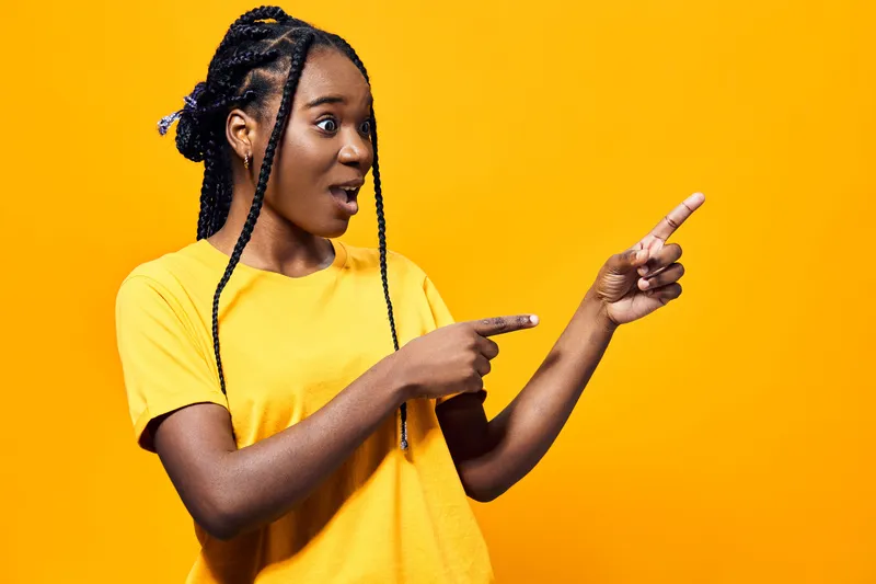 Excited young African American woman in a yellow shirt pointing at something surprising against a vibrant yellow background, conveying joy and enthusiasm