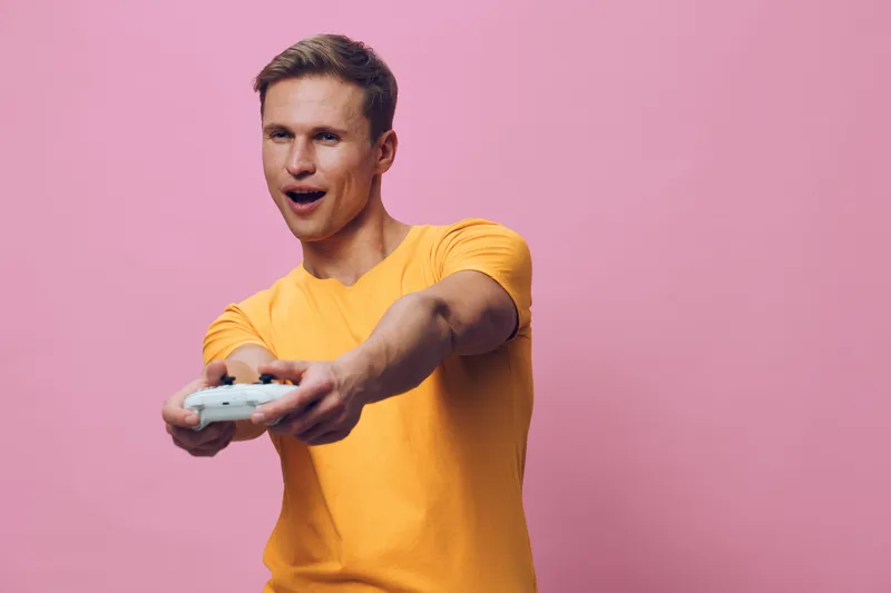 Excited young man playing video game holding controller wearing orange t-shirt isolated on pink background digital entertainment concept