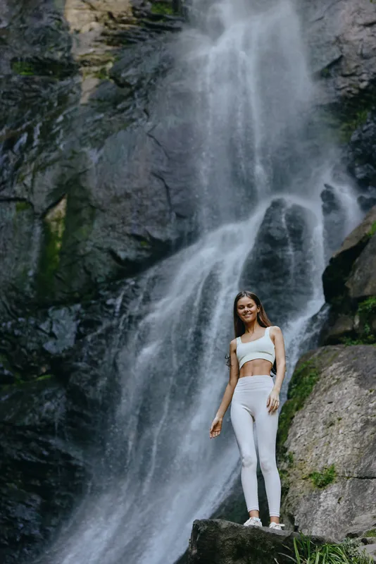 young woman standing by waterfall wearing white activewear, natural forest background, calm and peaceful atmosphere, outdoor fitness and nature