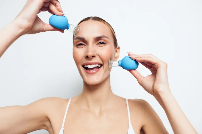 Woman performs facial massage with a blue tool, smiling against a white studio background, skincare and beauty routine for wellness and self care, emphasizing relaxation and bright skin