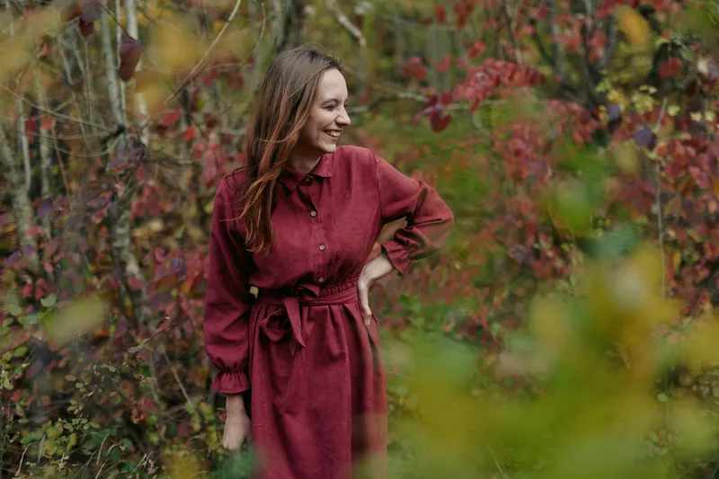 Candid autumn portrait of a smiling woman in a red dress among colorful foliage, conveying authentic warmth and genuine emotion in a believable, natural outdoor lifestyle scene.