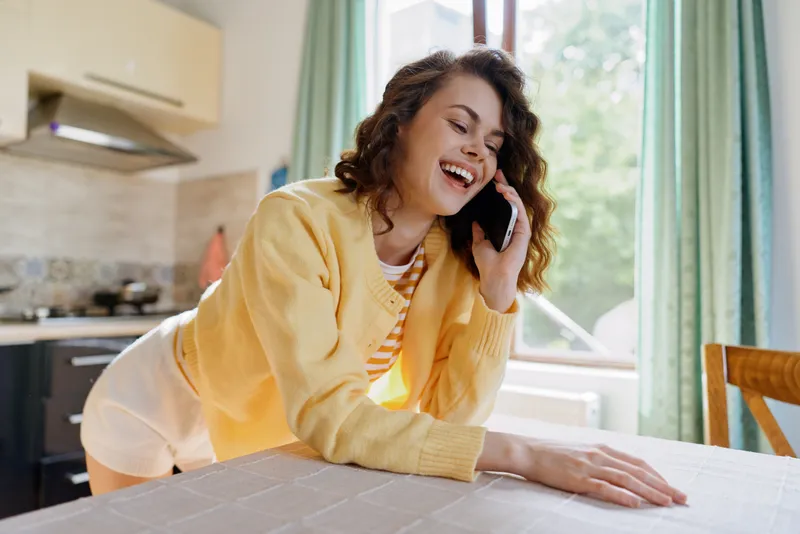 Smiling woman in a yellow cardigan leaning over a kitchen table while talking on a phone. Bright home setting conveys casual lifestyle, warmth, and everyday wellness.