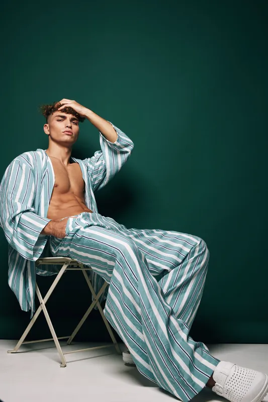 Young man in striped pajamas sitting on a chair with hand on forehead, relaxed pose and casual indoor fashion against dark green background in studio setting.