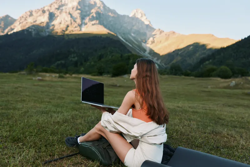 A woman sits in a sunlit meadow with a laptop, embracing nature while working outdoors. The peaceful landscape inspires focus and creative productivity in a bright setting.