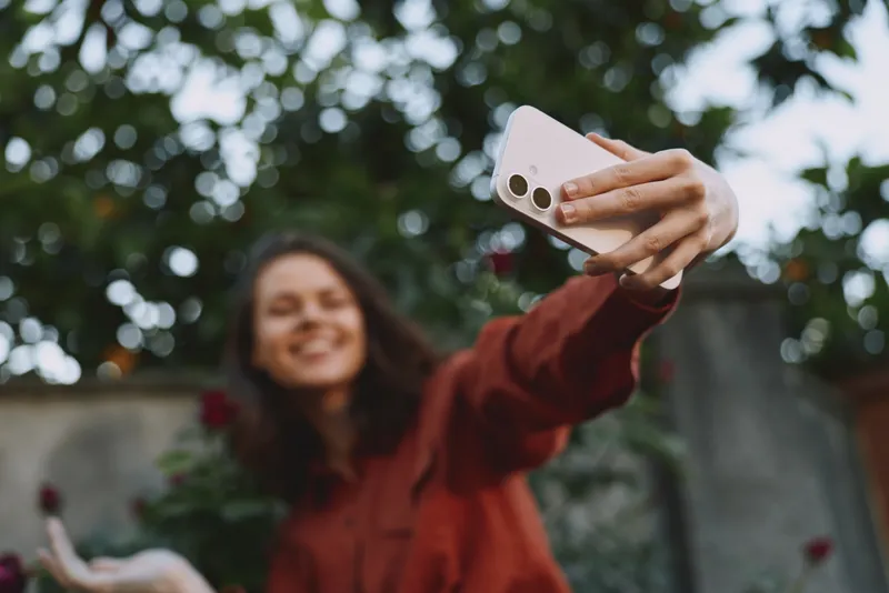 Smiling woman taking a selfie outdoors with a smartphone, blurred garden background. Casual fashion, lifestyle moment, playful mood, natural light, candid photography