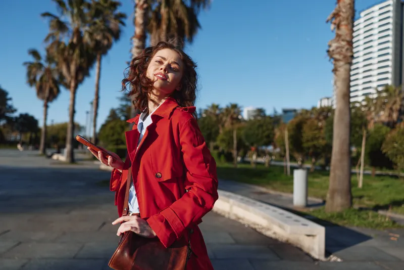 Young woman in red coat walking outdoors in sunny city park with palm trees, holding smartphone, casual style, confident expression, urban background