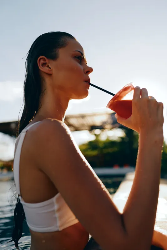 Young woman with wet hair drinks a refreshing red cocktail by the poolside in warm sunlight, enjoying a relaxed lifestyle moment on a summer day.