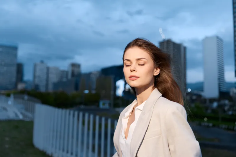 Woman in beige business suit with closed eyes in urban outdoor setting expressing calm and confidence during cloudy day, man, business, city, emotion, fashion