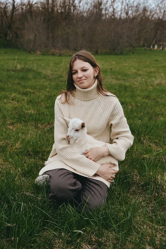 Young woman in a beige turtleneck sweater sits on grass holding a white baby goat in her arms during a calm outdoor moment in early spring.
