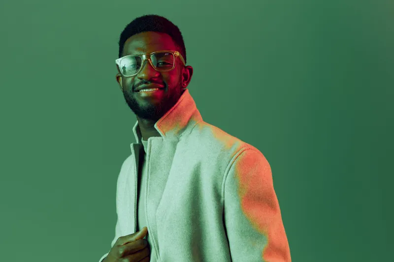 Stylish African American man in a light coat with glasses smiling against a green background, showcasing modern fashion and confidence