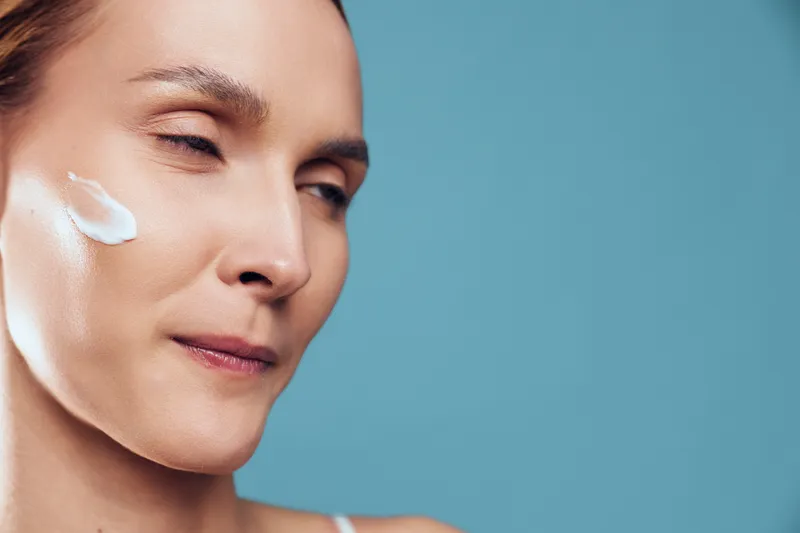 Close up of a smiling woman with cream on her cheek, calm expression, soft light, blue background, skincare moment