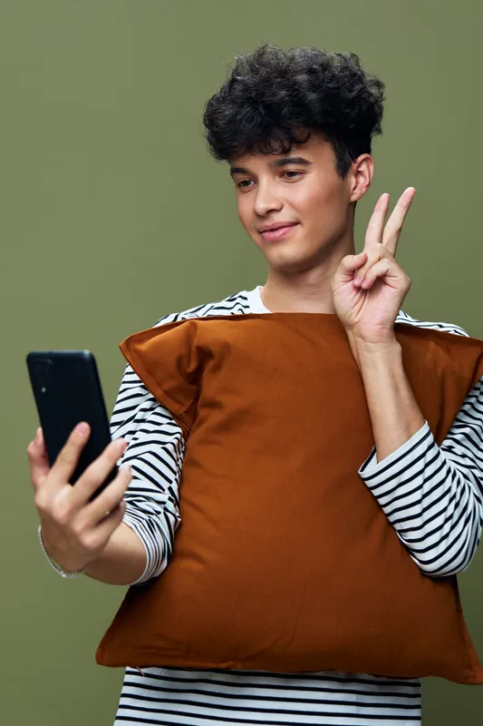 young man taking selfie holding brown pillow wearing striped shirt against green background, casual style, indoor lifestyle, happy expression