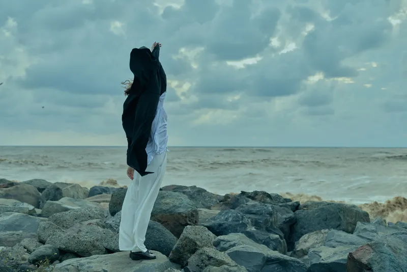 Woman standing on rocky shore under a cloudy sky, white dress and dark scarf flowing, contemplative scene of wind and ocean horizon