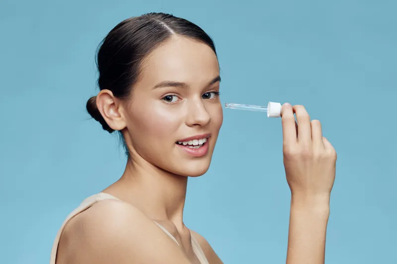 Young woman applying serum with dropper on a blue background, showcasing skincare routine, healthy skin, and beauty products