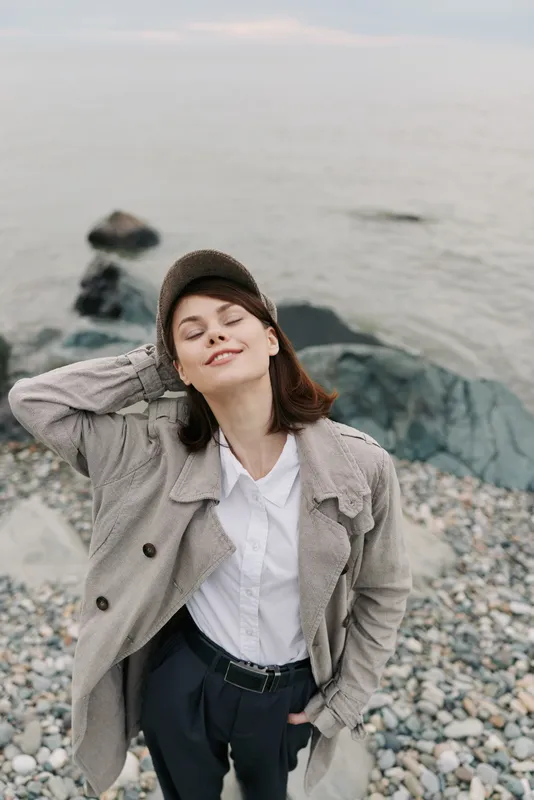 Young woman in casual outfit and cap stands on pebble beach near water, eyes closed and hand behind head, expressing calm and relaxation in natural coastal environment.