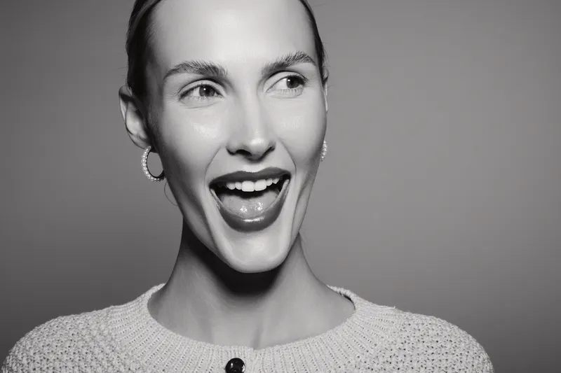 Happy Caucasian woman with light skin smiling widely wearing earrings and knitted sweater against grey studio background.