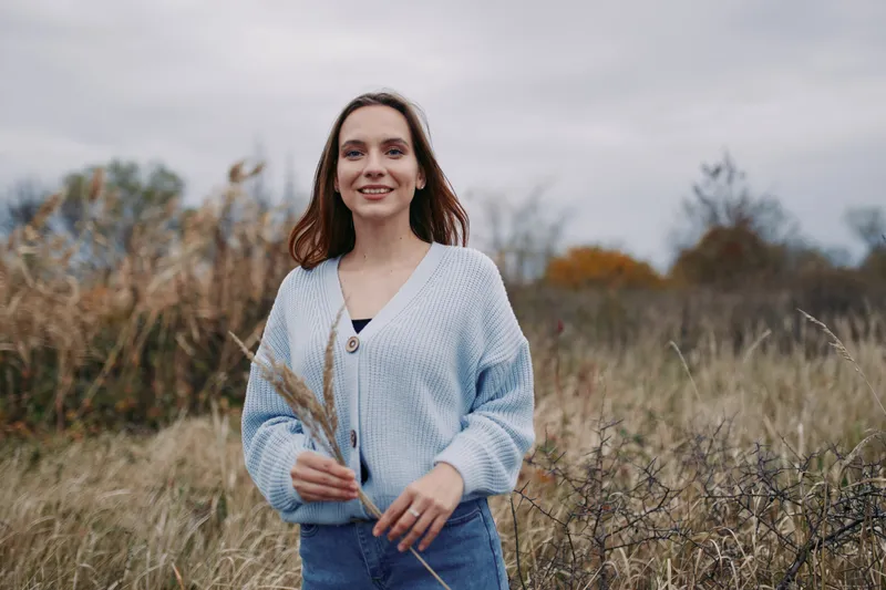 Candid portrait of a smiling woman standing in a dry grassy meadow under an overcast sky, natural light and subtle autumn tones convey authenticity and a believable everyday life moment.