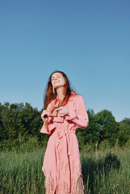 Young woman in a pink dress enjoying nature under a clear blue sky, exuding happiness and confidence in a lush green field