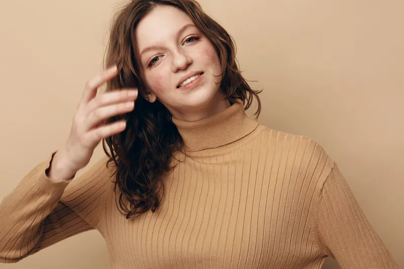 Young woman with curly hair wearing beige turtleneck sweater poses against beige background. Natural light highlights her relaxed expression and casual style. Concept of fashion and portrait.