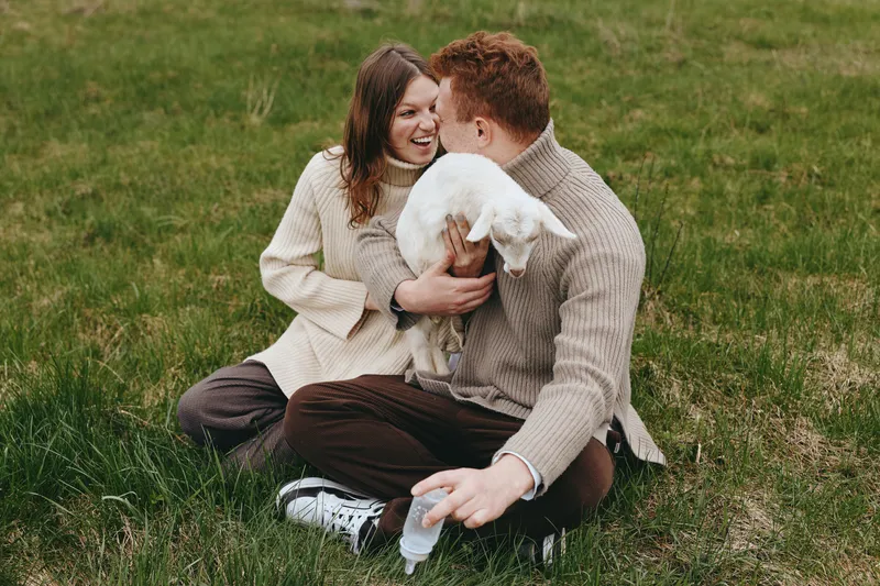 Happy couple on a grassy park, cuddling with their dog, smiling and relaxed, enjoying a sunny afternoon together outdoors, sharing warmth and genuine affection in a casual lifestyle moment.