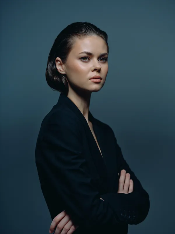 Serious woman in black jacket posing with arms crossed against dark background, showing confidence and professionalism.