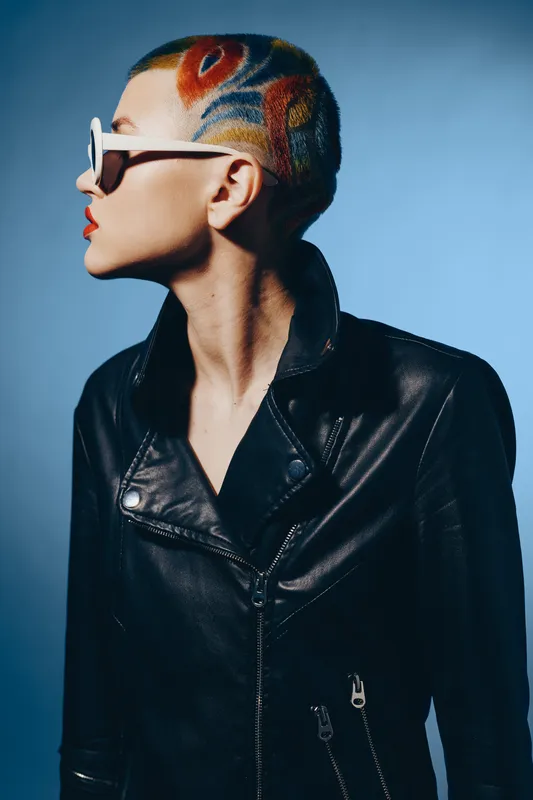 woman with colorful shaved hair and sunglasses wears a stylish black leather jacket against blue background, showcasing edgy fashion and bold style.