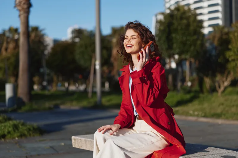 A young woman in a red coat sits on a concrete bench in an urban park, talking on a phone and smiling, capturing a candid moment of everyday urban life.
