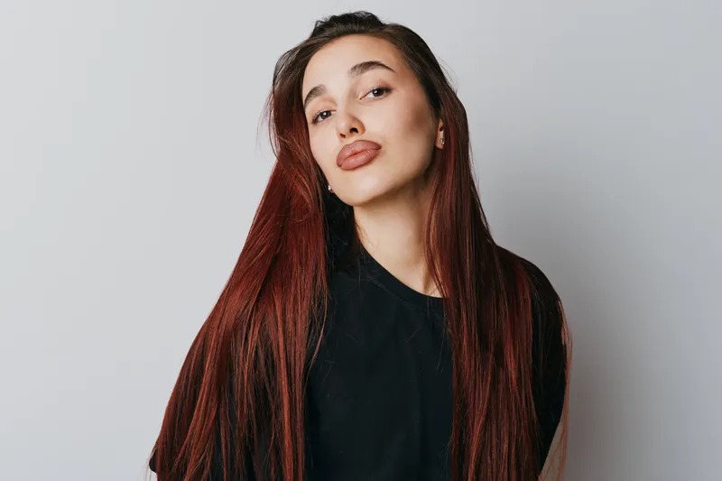 Young woman with long red hair and natural makeup posing confidently against plain background. Casual style with black top and relaxed expression for portrait photography.