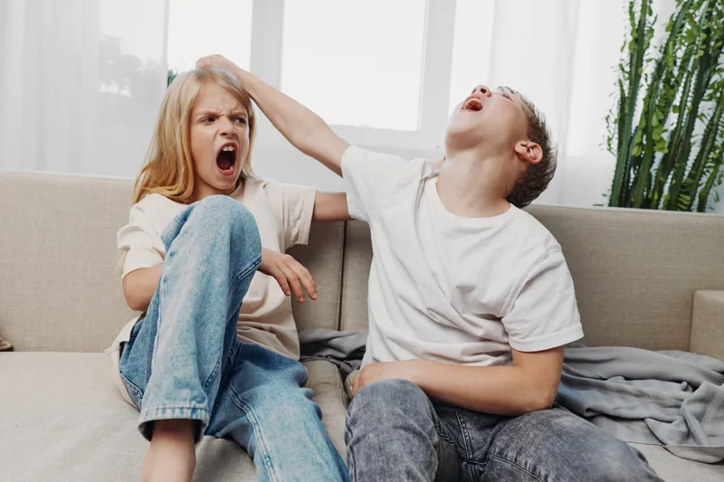 Two children in casual clothes sit on a couch indoors, one boy playfully pulling a girl’s hair while she reacts with a frustrated expression. Concept of sibling rivalry and childhood conflict.
