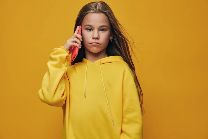 Girl with long hair in yellow hoodie talking on red smartphone against yellow background, expression serious, communication and technology concept.