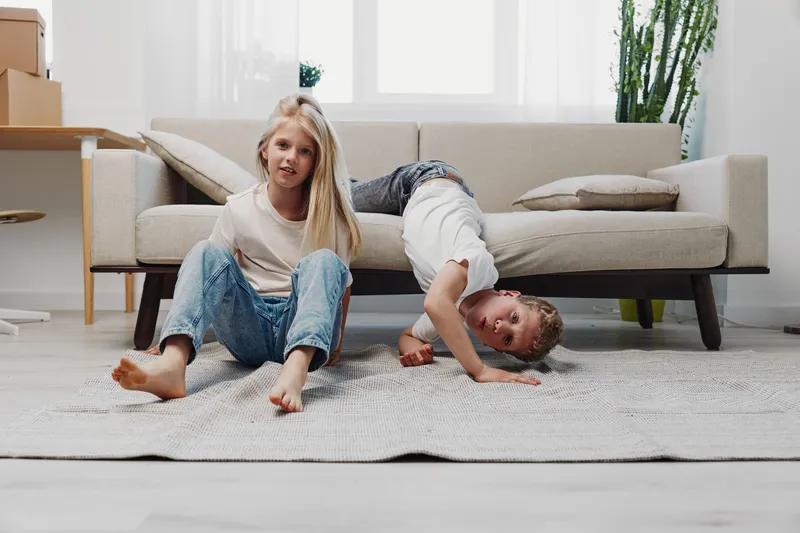 Two children playing on carpet in bright living room, one girl sitting and one boy bending down near sofa, casual home activity concept.