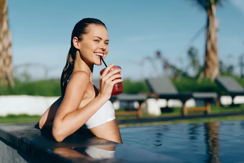 A relaxed woman in a white swimsuit sits at pool edge, sipping a red drink, sunlight on skin, bright outdoor scene, conveying lifestyle, leisure, wellness, and summer mood for bright mood.