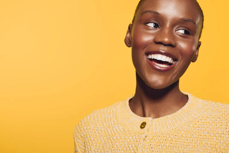Happy smiling African woman with dark skin wearing yellow sweater looks away on yellow background. Concept of positive emotions, beauty, fashion, and natural skin glow in studio lighting.