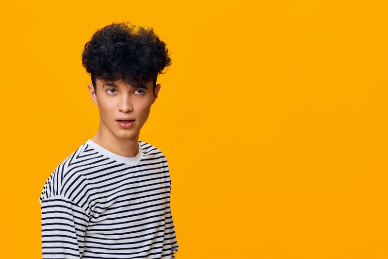 Young man with curly hair wearing a black and white striped shirt posing against a vibrant orange background with a surprised facial expression, casual style.