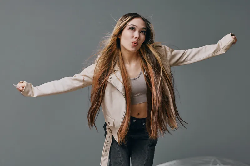 Young woman with long braided hair and beige jacket dancing and posing energetically against gray background. Casual clothing and expressive facial expression with motion.