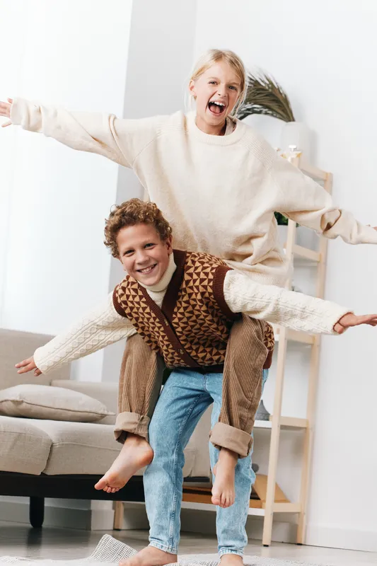 Two joyful children playfully jump indoors, sharing laughter as they ride on a couch. Spontaneous moment captures energy, warmth, and friendly cheer in a cozy home setting.