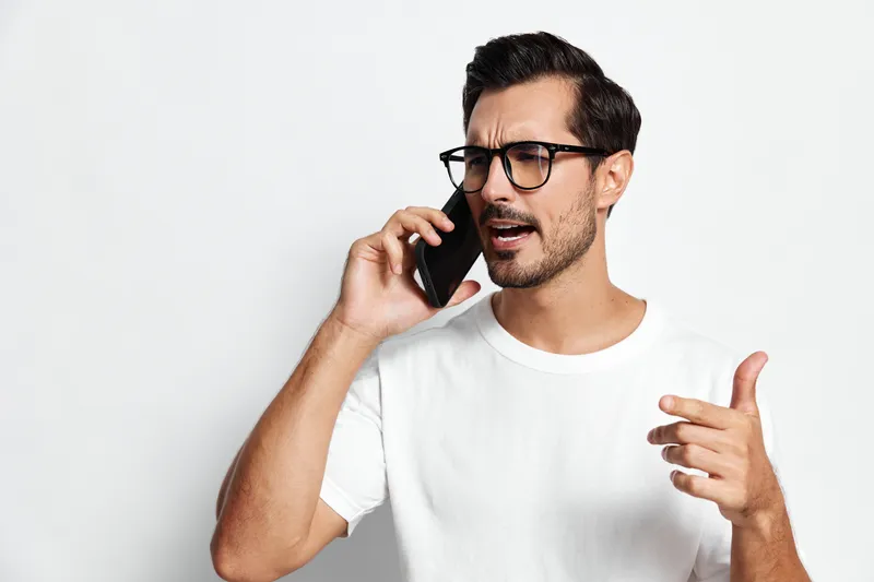 Full body frustrated young caucasian man wearing glasses talking on mobile phone with serious expression isolated on plain solid white background. People lifestyle concept.
