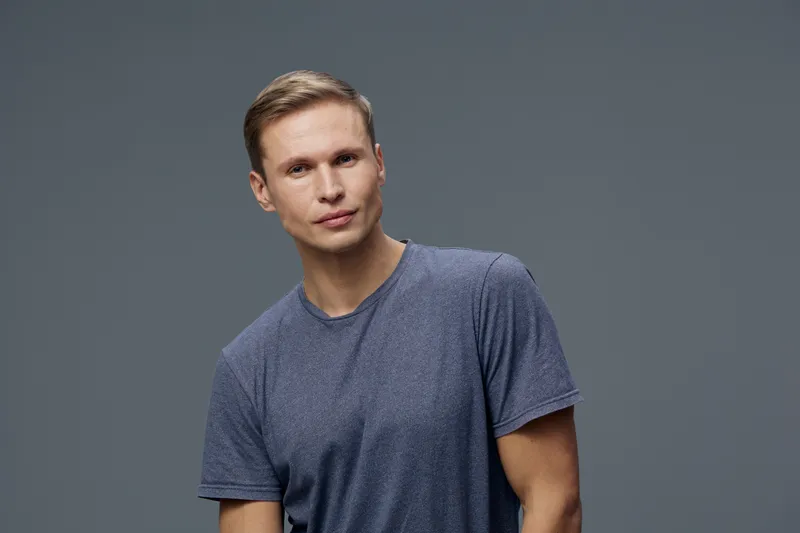 Young man with short brown hair and fair skin wearing casual blue t-shirt looking confidently at camera isolated on plain gray background in studio portrait. People lifestyle concept, male fashion.