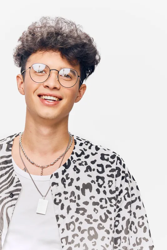 young man wearing round glasses and trendy leopard print shirt smiling with curly hair against a white background in casual style